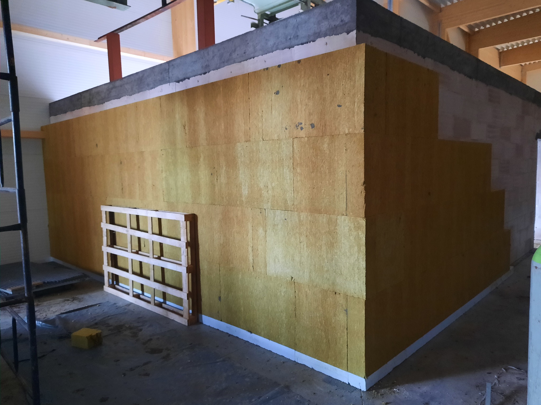 Ocieplenie ścian wełną mineralną w surowym stanie, widoczna drabina i paleta drewniana, etap prac wykończeniowych.