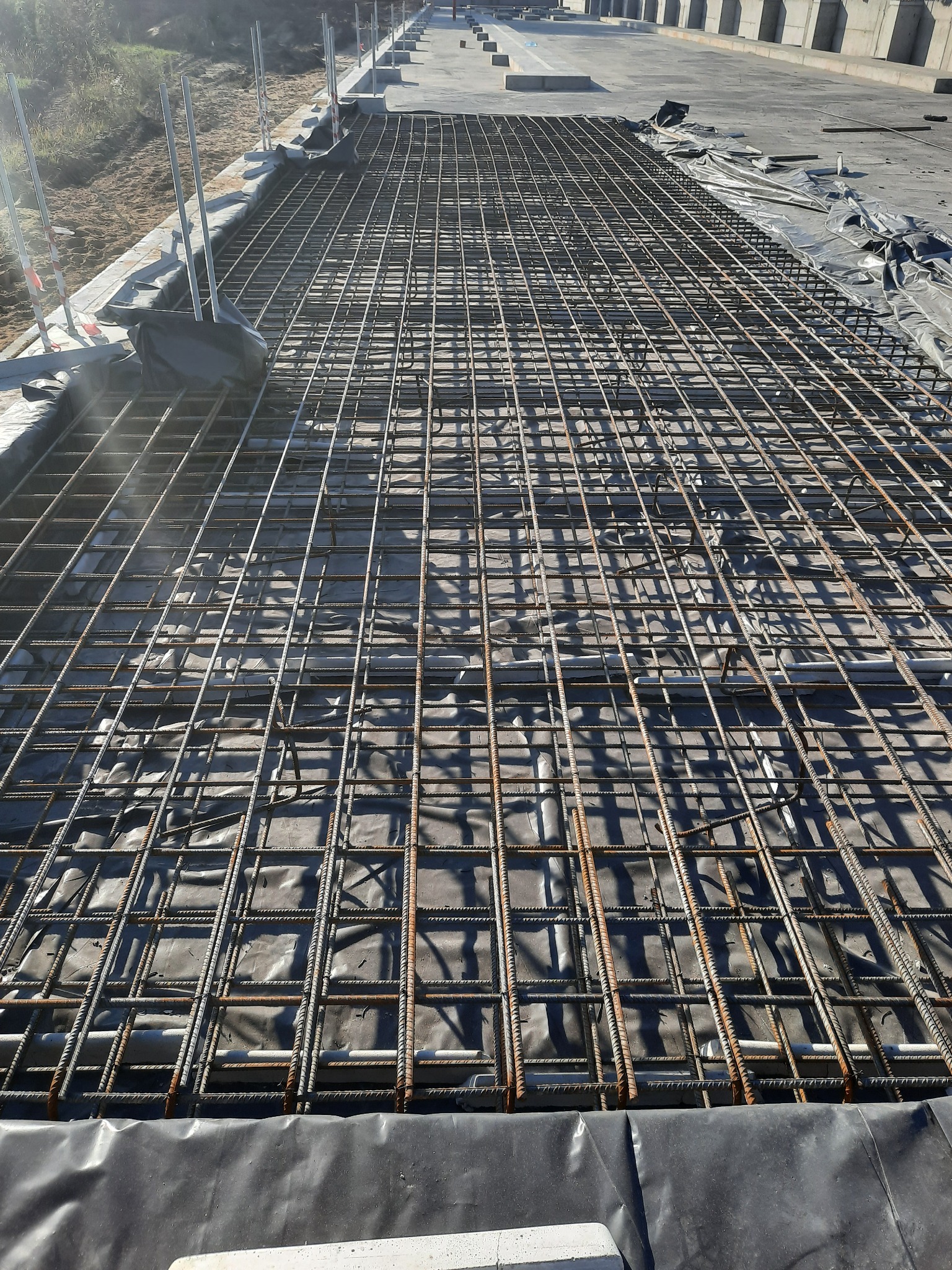 Zbrojenie płyty fundamentowej z siatki stalowej na czarnej folii izolacyjnej, przygotowane do zalania betonem, widoczne elementy szalunku i pionowe słupki.