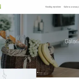 Kosz piknikowy z winem, pieczywem, bananami i winogronami na stole w jasnym, nowoczesnym wnętrzu, widok z poziomu blatu, strona internetowa korbtex.pl w tle.