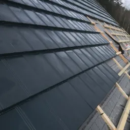 Montaż grafitowej dachówki na łatach z widoczną membraną dachową i fragmentem lasu w tle.