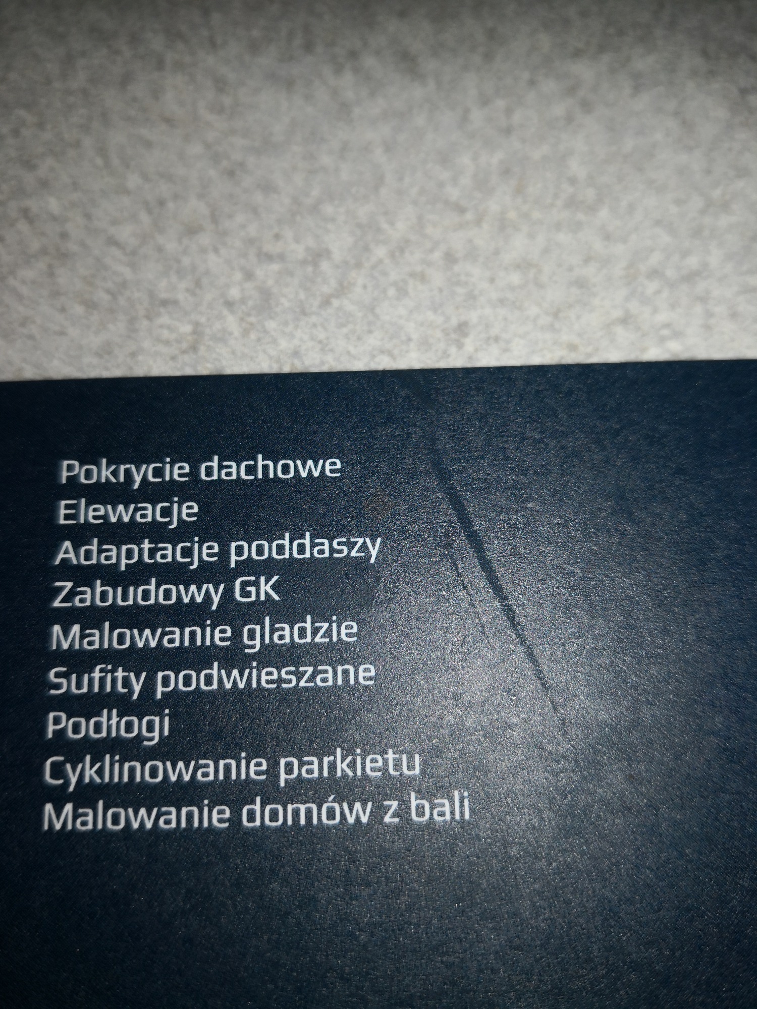 Zbliżenie na fragment materiału reklamowego z listą usług budowlanych, w tym pokrycie dachowe, elewacje, adaptacje poddaszy, zabudowy GK, malowanie gładzi, sufity podwieszane, podłogi...