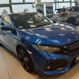 Niebieski samochód Honda Civic na tle salonu samochodowego z widocznymi plakatami reklamowymi i innym modelem Hondy HR-V
