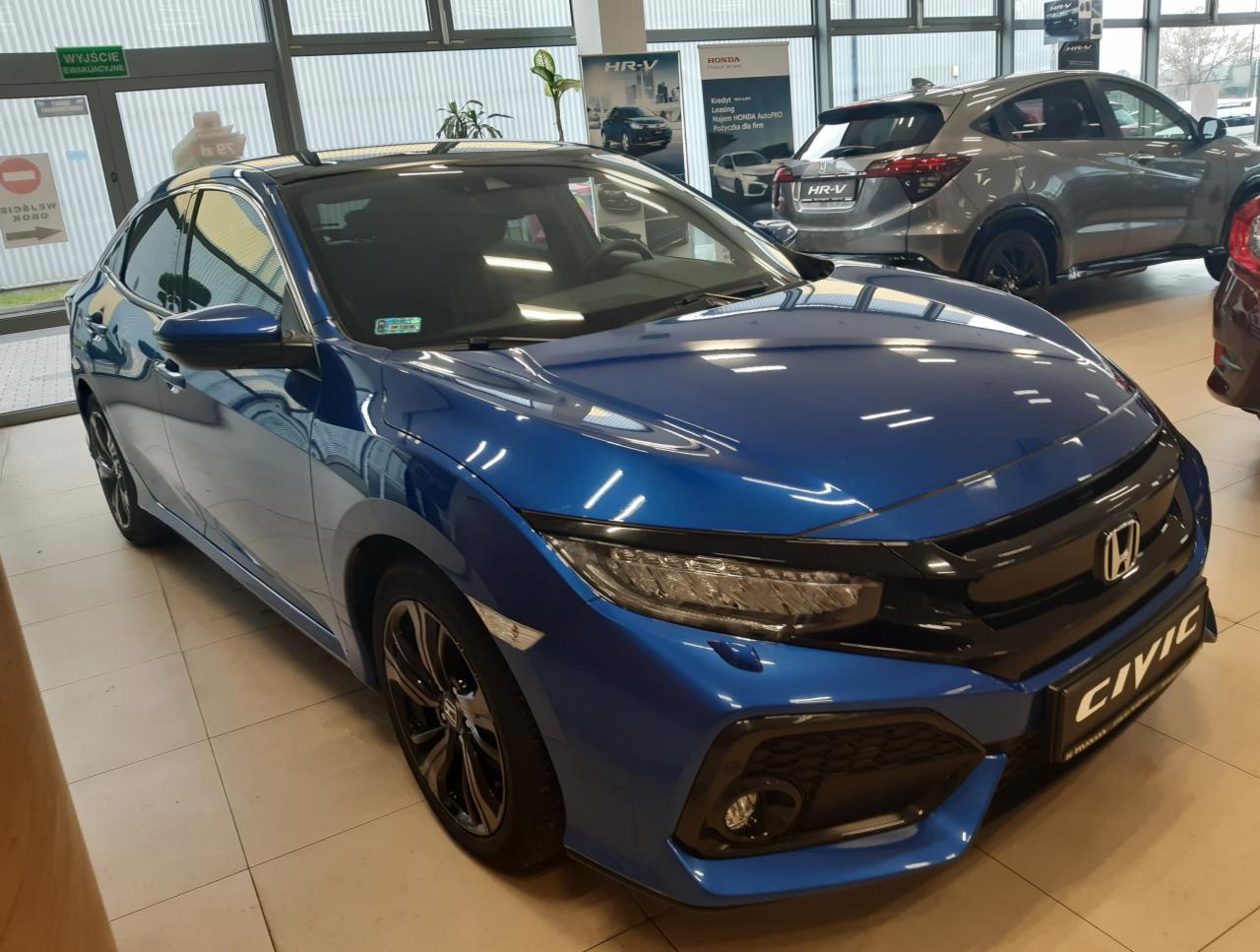Niebieski samochód Honda Civic na tle salonu samochodowego z widocznymi plakatami reklamowymi i innym modelem Hondy HR-V