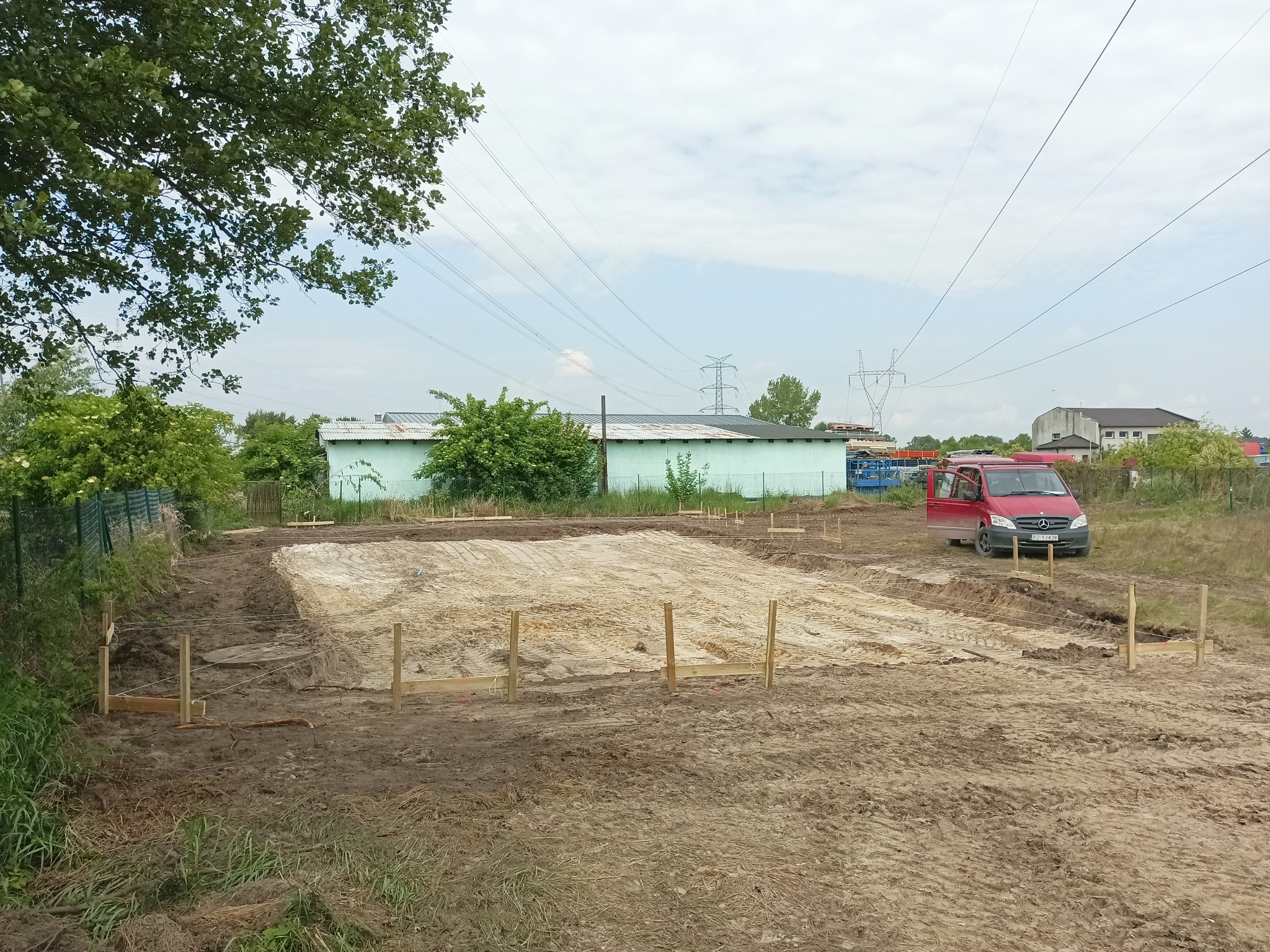 Przygotowany teren pod budowę domu z wytyczonym obrysem fundamentów, drewniane słupki i sznurki, czerwony samochód dostawczy w tle, pochmurne niebo.