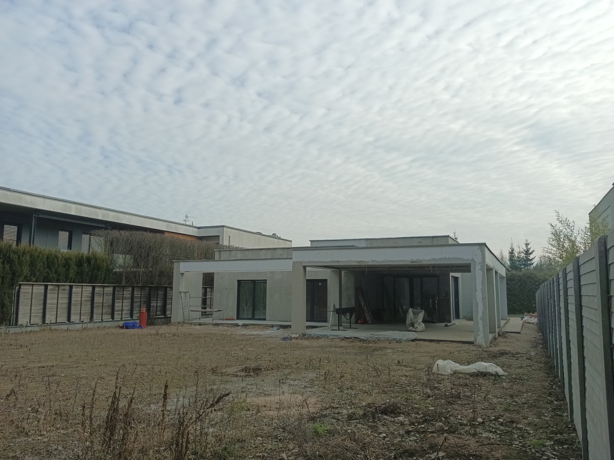 Surowy, parterowy dom z płaskim dachem w trakcie budowy. Widoczne niezadaszone patio, okna i drzwi balkonowe. Na pierwszym planie niezagospodarowany teren.