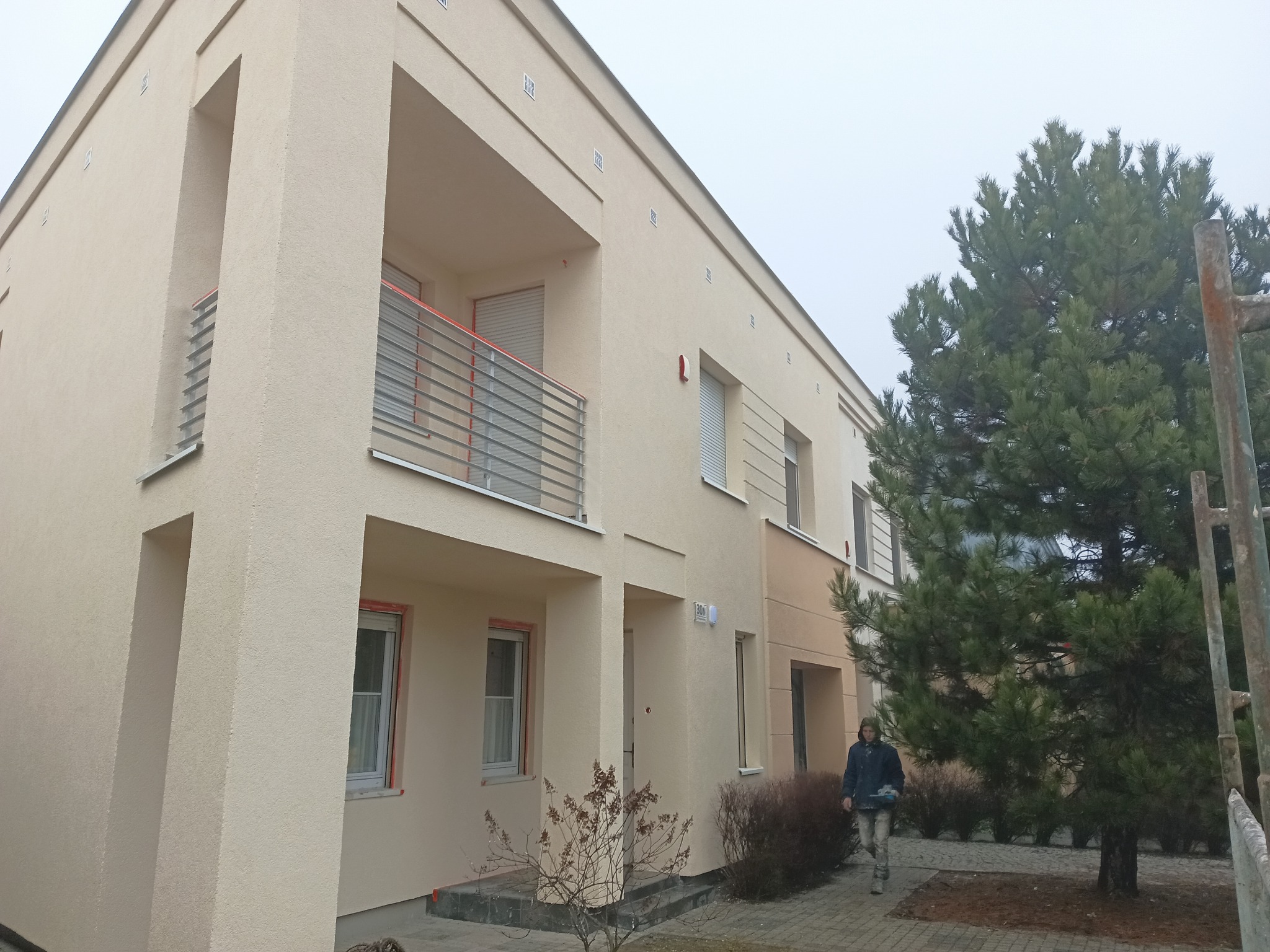 Dwukondygnacyjny dom z balkonem i beżową elewacją, widok z zewnątrz. Osoba przechodzi obok budynku. Obok rośnie duża sosna. Pochmurne niebo w tle.