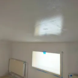 Świeżo pomalowane na biało wnętrze pokoju z zabezpieczonym oklejonymi taśmą okiennicami i grzejnikami, widoczne niedociągnięcia i mokre plamy na suficie.