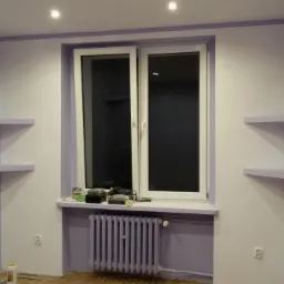 Pokój po odświeżeniu w Zielonej Górze: ściany pomalowane na biało i liliowo, z liliowymi półkami narożnymi, okno z białymi ramami nad liliowym grzejnikiem.