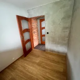 Pokój po odświeżeniu: parkiet laminowany, szara ściana z efektem dekoracyjnym, otwarte drewniane drzwi z przeszkleniami, widoczna ceglana ściana w przejściu do kolejnego pomieszczenia.