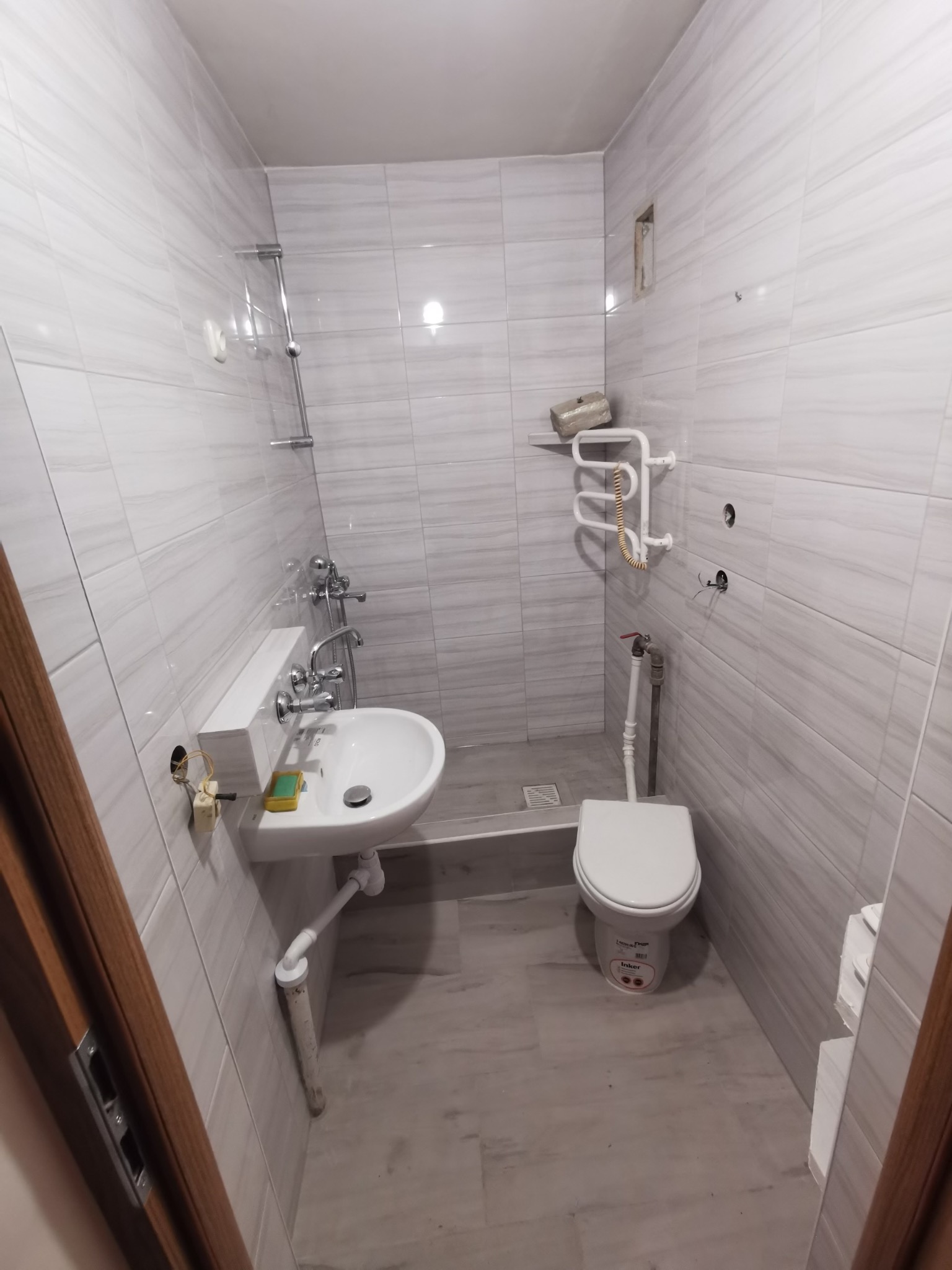 Niewielka łazienka z szarymi płytkami, umywalką z baterią, toaletą i grzejnikiem drabinkowym, widoczne niedokończone podłączenia elektryczne i hydrauliczne.