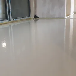 Świeżo wylana, gładka, jasnoszara posadzka w pomieszczeniu w stanie surowym, zabezpieczona folią przy ścianach i oknach marki VEKA.