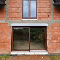 ST SITO PROJECT SPÓŁKA Z OGRANICZONĄ ODPOWIEDZIALNOŚCIĄ - Dwa brązowe okna w surowej, czerwonej ceglanej ścianie budynku. Dolne okno to przesuwne, górne z roletą. Widoczne detale montażu i otoczenie budynku.