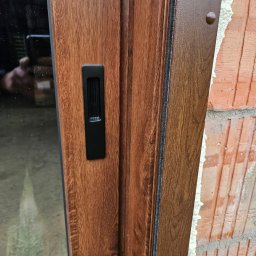 ST SITO PROJECT SPÓŁKA Z OGRANICZONĄ ODPOWIEDZIALNOŚCIĄ - Brązowe drzwi przesuwne z czarnym uchwytem, widoczne na tle surowej ściany z cegły i pianki montażowej. Detal wykończenia i montażu.