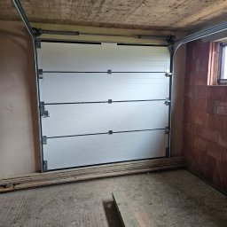 ST SITO PROJECT SPÓŁKA Z OGRANICZONĄ ODPOWIEDZIALNOŚCIĄ - Zamknięta, segmentowa brama garażowa w surowym wnętrzu z betonowym sufitem i ścianami z cegły. Widoczne prowadnice i sprężyny bramy. Na podłodze leżą deski.