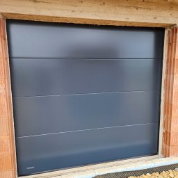 ST SITO PROJECT SPÓŁKA Z OGRANICZONĄ ODPOWIEDZIALNOŚCIĄ - Zamknięta, segmentowa brama garażowa w kolorze grafitowym, zamontowana w surowym otworze budynku z cegły. Widoczne detale montażu i otoczenie budowy.