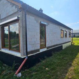 ST SITO PROJECT SPÓŁKA Z OGRANICZONĄ ODPOWIEDZIALNOŚCIĄ - Surowy budynek z brązowymi oknami PCV i zamontowanymi roletami zewnętrznymi. Widoczne detale montażu i ocieplenia fundamentów. Budowa w toku, zielona trawa.