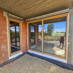 ST SITO PROJECT SPÓŁKA Z OGRANICZONĄ ODPOWIEDZIALNOŚCIĄ - Montaż dużych, przesuwnych okien z drewnianą ramą w surowym stanie budynku. Widoczne cegły i beton. Na zewnątrz drabina i odbicie osoby w szybie.