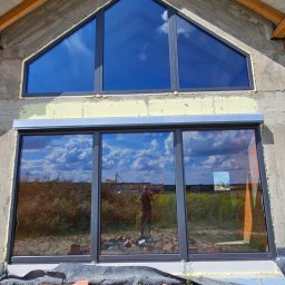 ST SITO PROJECT SPÓŁKA Z OGRANICZONĄ ODPOWIEDZIALNOŚCIĄ - Nowe okna PCV w surowym budynku z szarą elewacją. Trójkątne okno na górze i duże okno na dole z roletą. Widoczne odbicie fotografa w oknach.