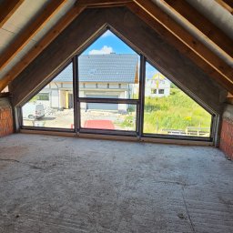 oknatychy.pl ST SITO PROJECT Sp. z o.o. oknatychy.pl - Surowy strych z oknami o nietypowym kształcie, widok na domy i zieleń. Okna z czarnymi ramami, surowy beton i cegła. Drewniana konstrukcja dachu.