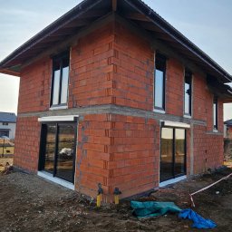 ST SITO PROJECT SPÓŁKA Z OGRANICZONĄ ODPOWIEDZIALNOŚCIĄ - Surowy budynek z czerwonej cegły z zamontowanymi oknami i drzwiami przesuwnymi w czarnych ramach. Widoczne elementy instalacji na zewnątrz budynku.