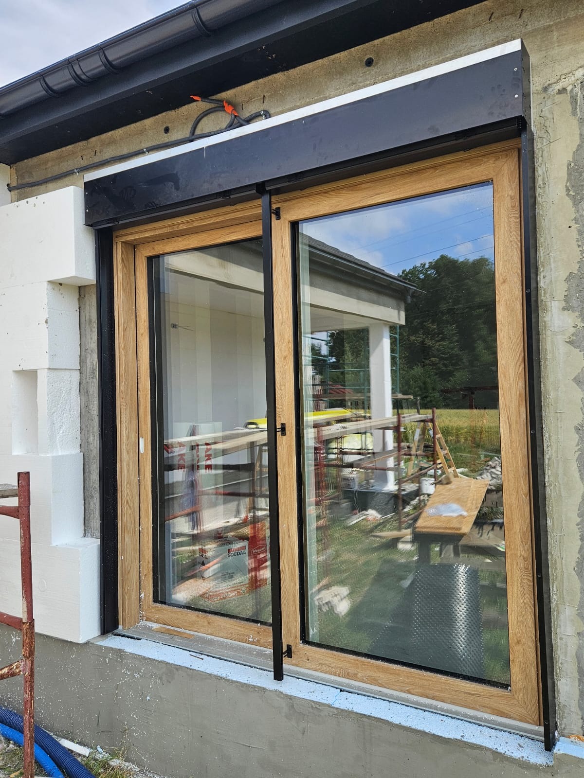 Zamontowane przesuwne okno z roletą zewnętrzną w elewacji budynku w trakcie prac wykończeniowych. Widoczne rusztowania i materiały budowlane.