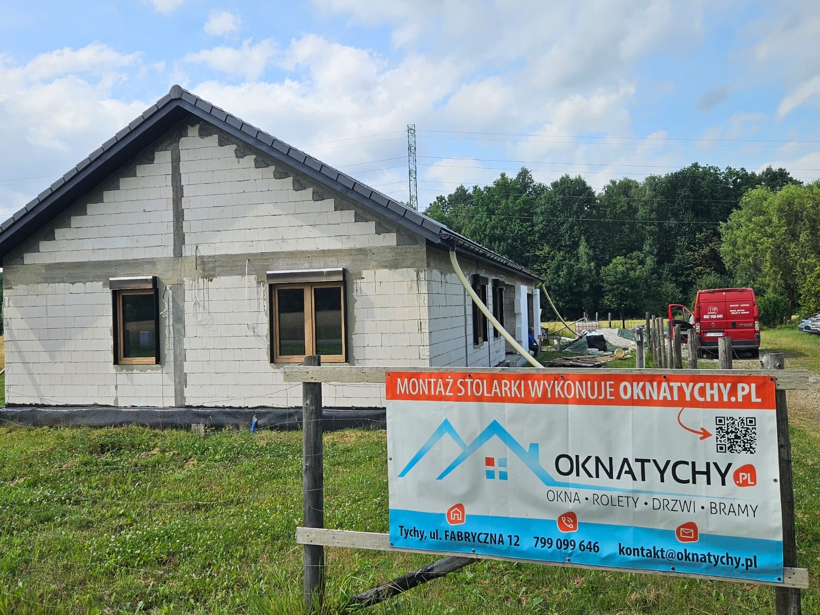 Dom w trakcie budowy z zamontowanymi oknami. Na pierwszym planie baner reklamowy firmy oferującej okna, rolety, drzwi i bramy. W tle las i błękitne niebo.