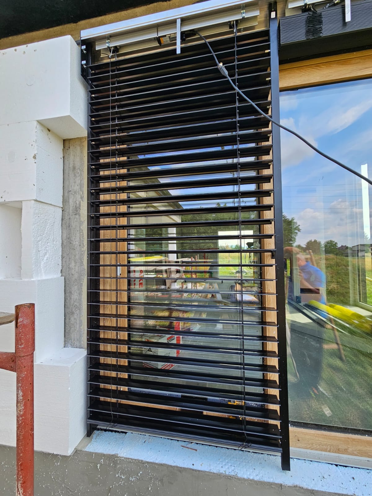 Zamontowane czarne żaluzje fasadowe zewnętrzne przy oknie z drewnianą ramą. Widoczne białe bloczki konstrukcyjne i częściowo odbicie osoby w szybie.