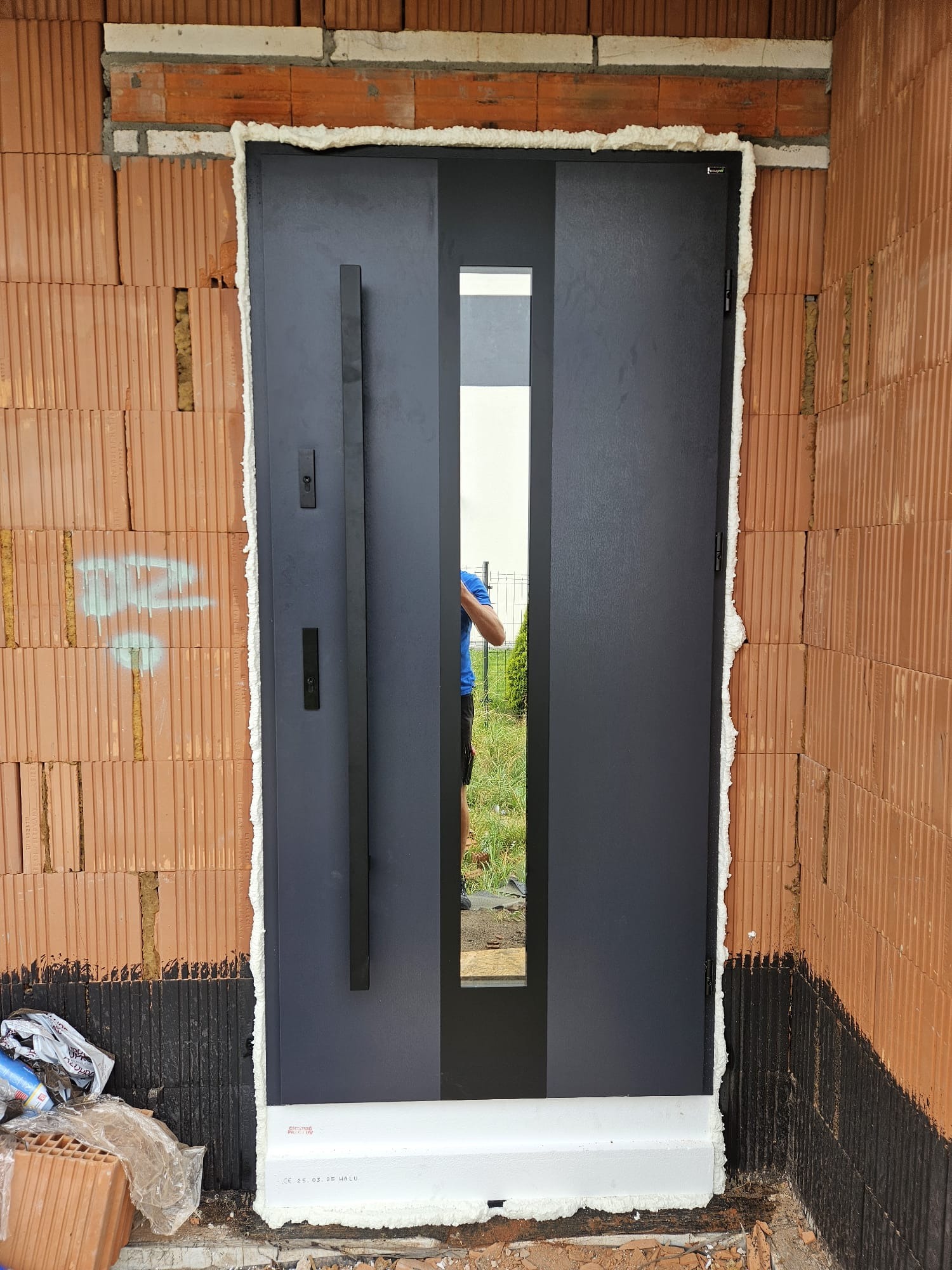 Ciemne drzwi wejściowe z szybą, zamontowane w surowym murze z cegły, uszczelnione pianką montażową. Widoczny fragment osoby w odbiciu szyby.