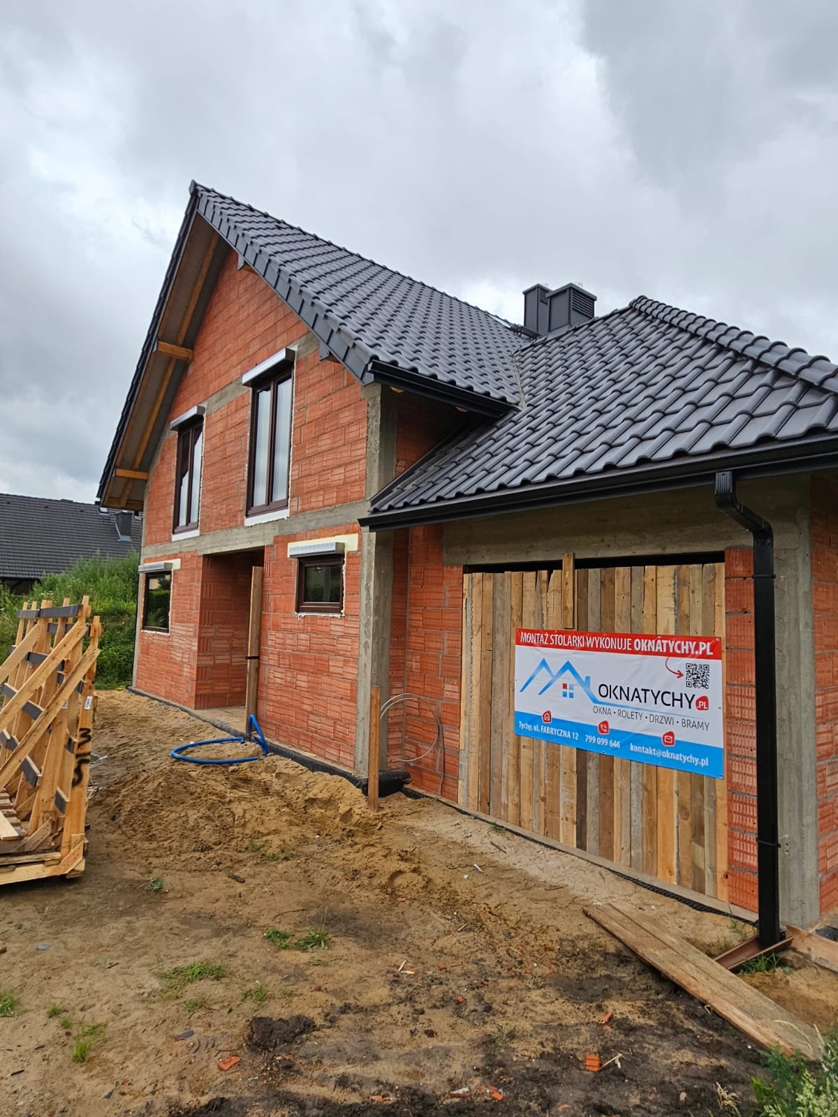 Surowy budynek z cegły z zamontowanymi oknami i tymczasową bramą garażową z desek. Widoczny baner reklamowy firmy stolarskiej. Dach pokryty czarną dachówką.