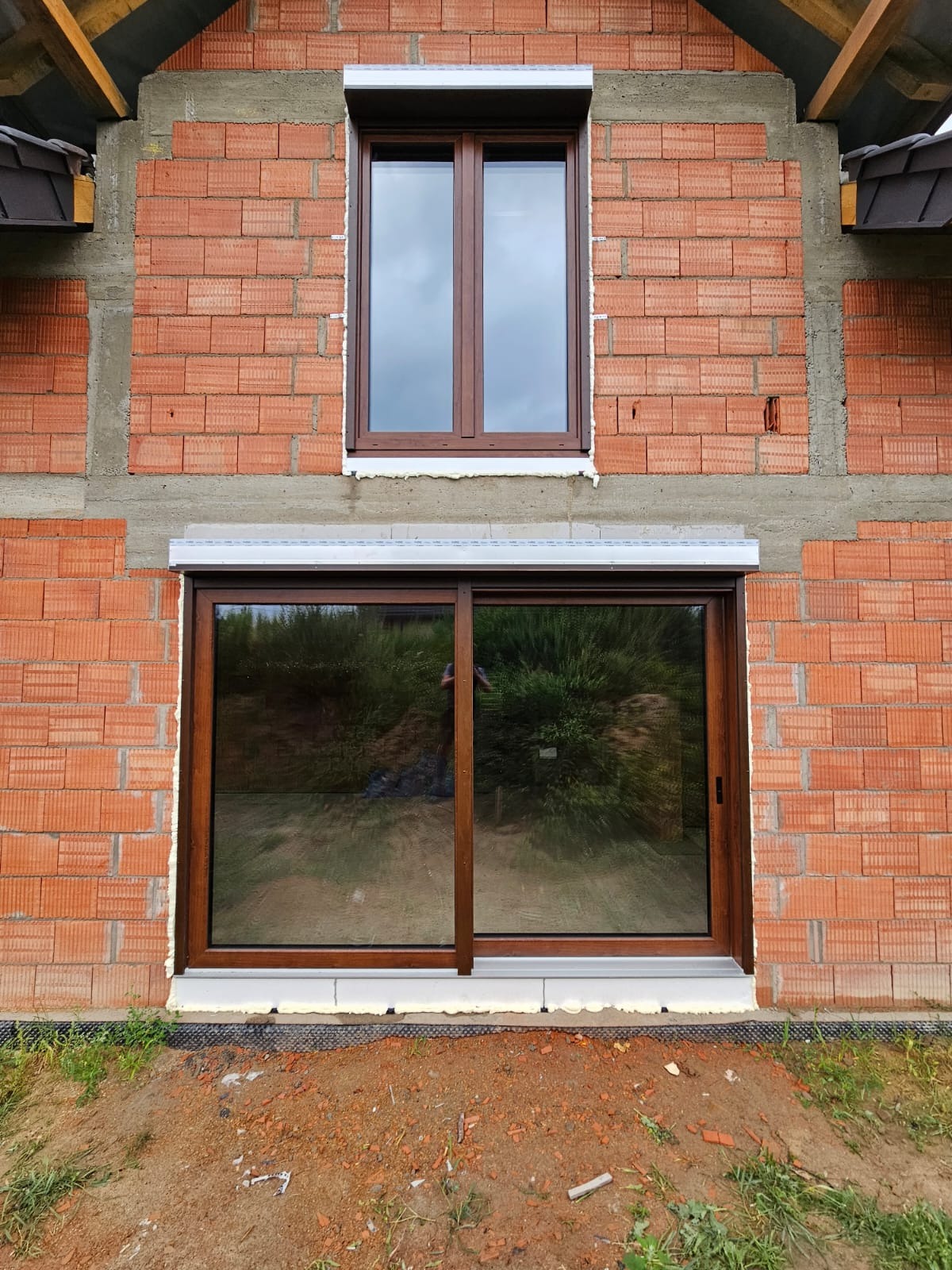 Dwa brązowe okna w surowej, czerwonej ceglanej ścianie budynku. Dolne okno to przesuwne, górne z roletą. Widoczne detale montażu i otoczenie budynku.