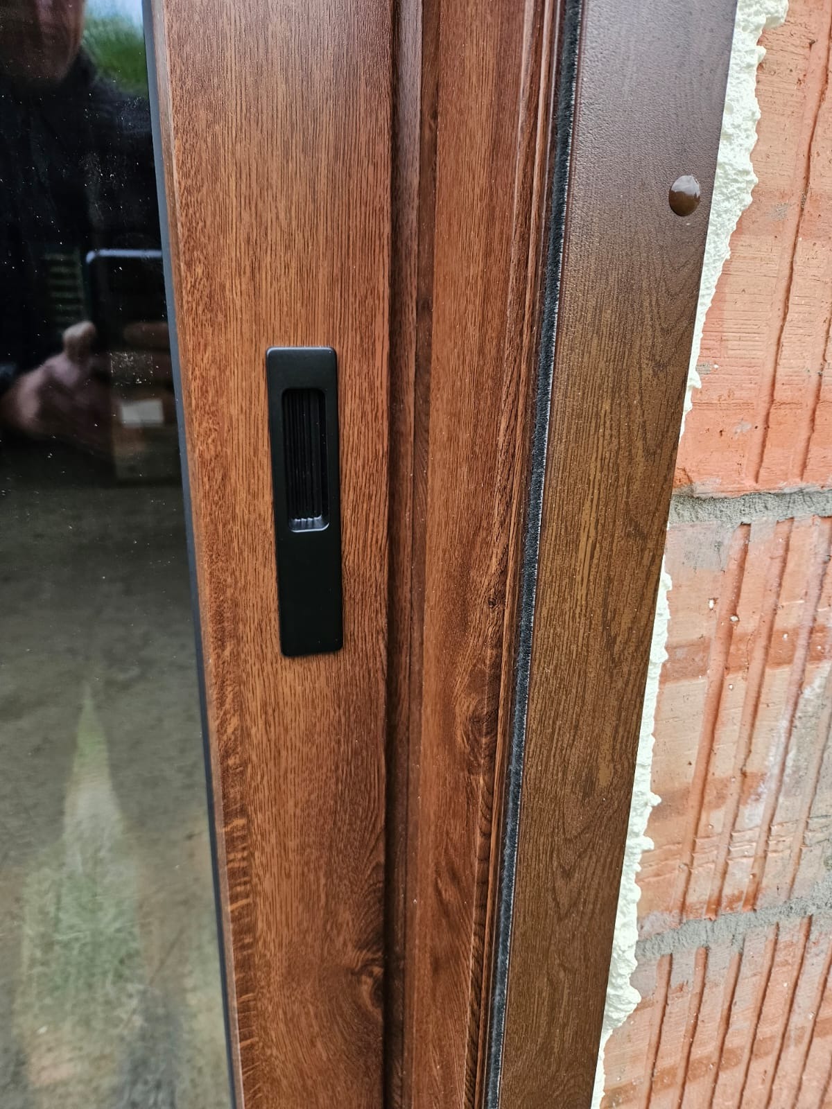 Brązowe drzwi przesuwne z czarnym uchwytem, widoczne na tle surowej ściany z cegły i pianki montażowej. Detal wykończenia i montażu.