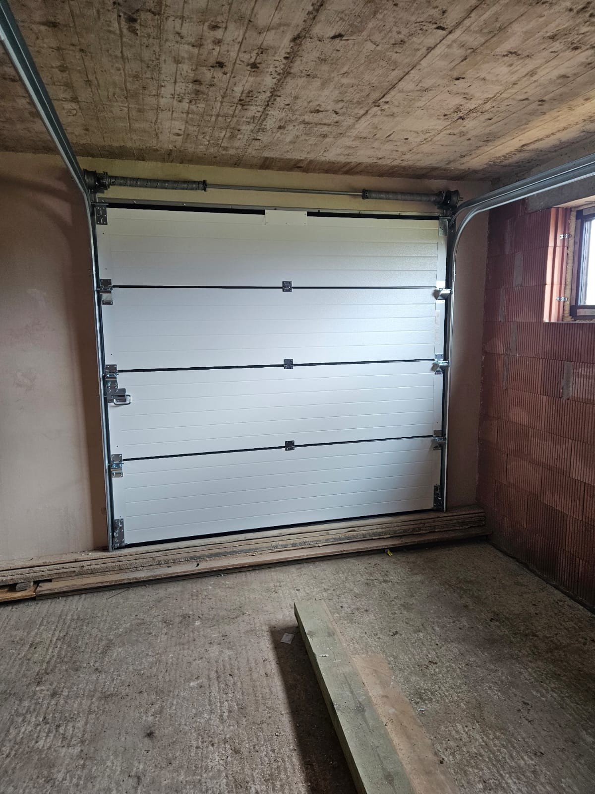Zamknięta, segmentowa brama garażowa w surowym wnętrzu z betonowym sufitem i ścianami z cegły. Widoczne prowadnice i sprężyny bramy. Na podłodze leżą deski.