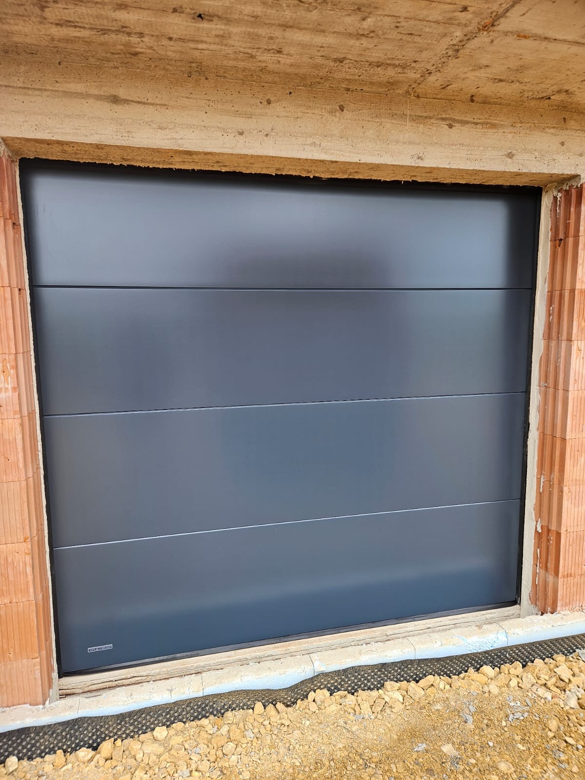 Zamknięta, segmentowa brama garażowa w kolorze grafitowym, zamontowana w surowym otworze budynku z cegły. Widoczne detale montażu i otoczenie budowy.