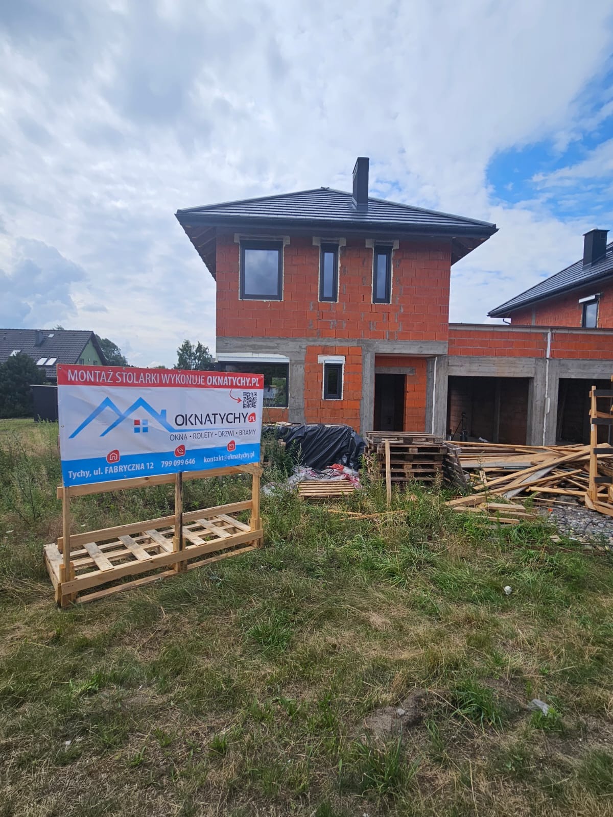 Dom w trakcie budowy z zamontowanymi oknami, widoczny baner reklamowy firmy OKNATYCHY.PL oferującej okna, rolety, drzwi i bramy. Palety i deski na trawie.