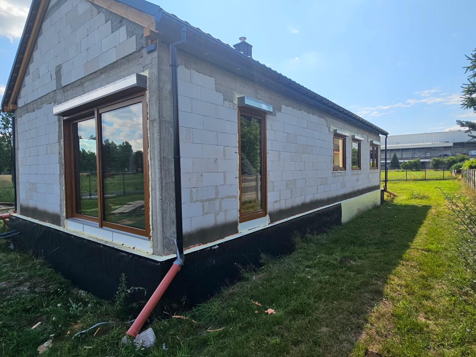Surowy budynek z brązowymi oknami PCV i zamontowanymi roletami zewnętrznymi. Widoczne detale montażu i ocieplenia fundamentów. Budowa w toku, zielona trawa.
