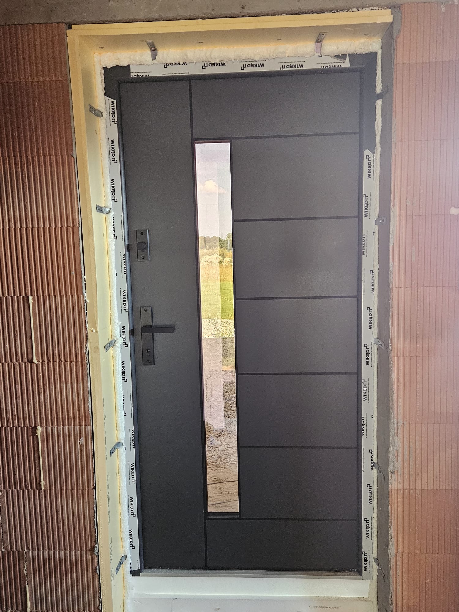Nowoczesne, grafitowe drzwi wejściowe z przeszkleniem, zamontowane w surowym murze. Widoczne detale montażu i uszczelnienia. Minimalistyczny design.