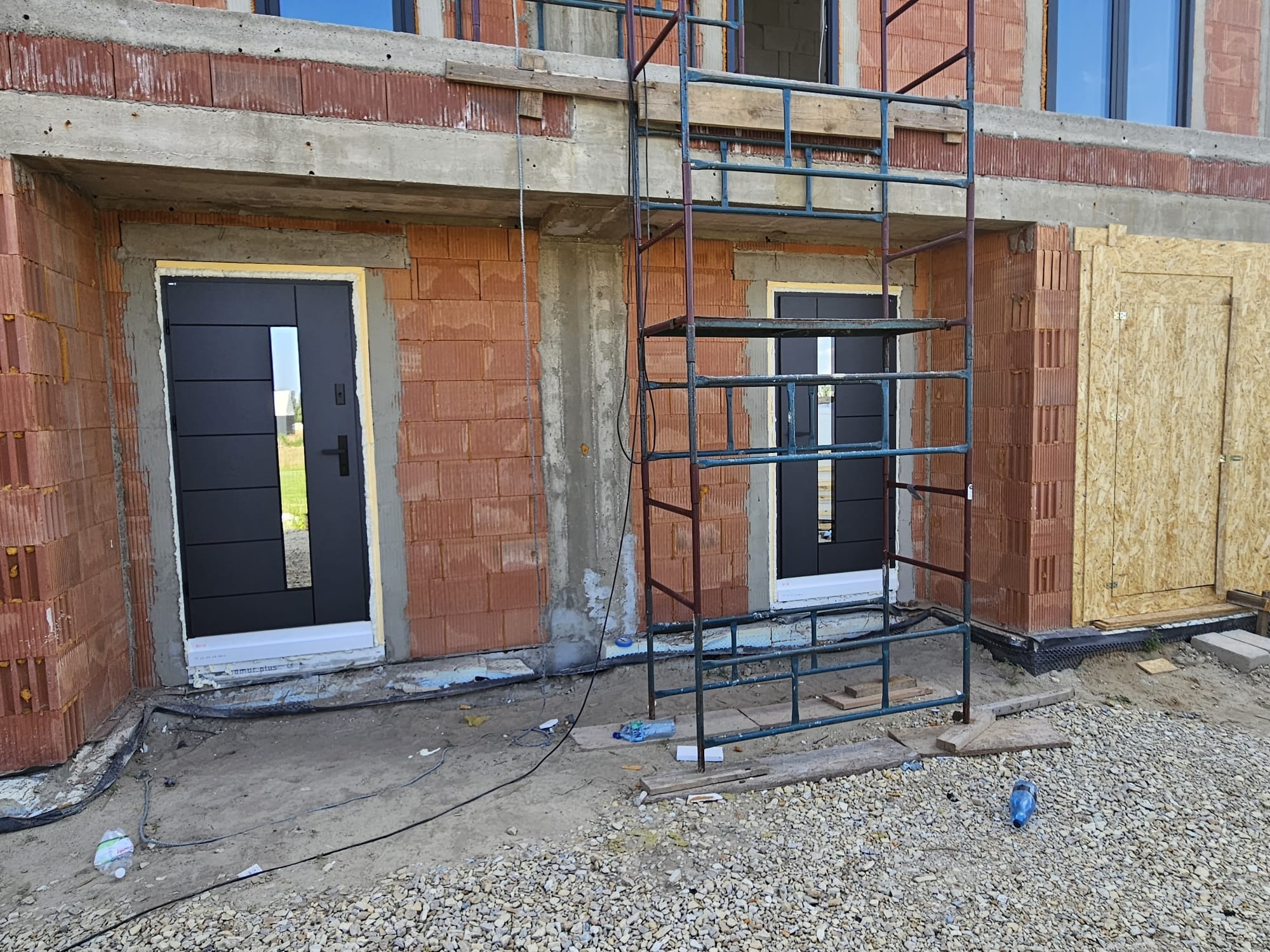 Montaż czarnych drzwi zewnętrznych z szybą w surowym budynku z czerwonej cegły. Widoczne rusztowanie i gruz.