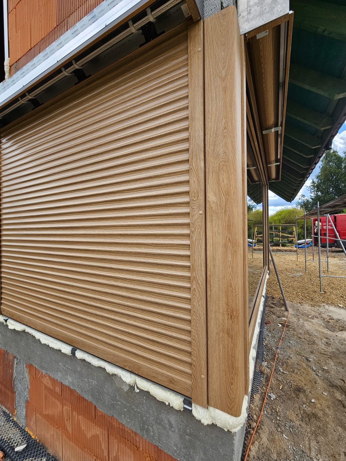 Zamknięta roleta zewnętrzna w kolorze drewna, zamontowana na oknie w nowym budynku. Widoczne detale montażu i faktura rolety. Ujęcie z dołu, z boku budynku.