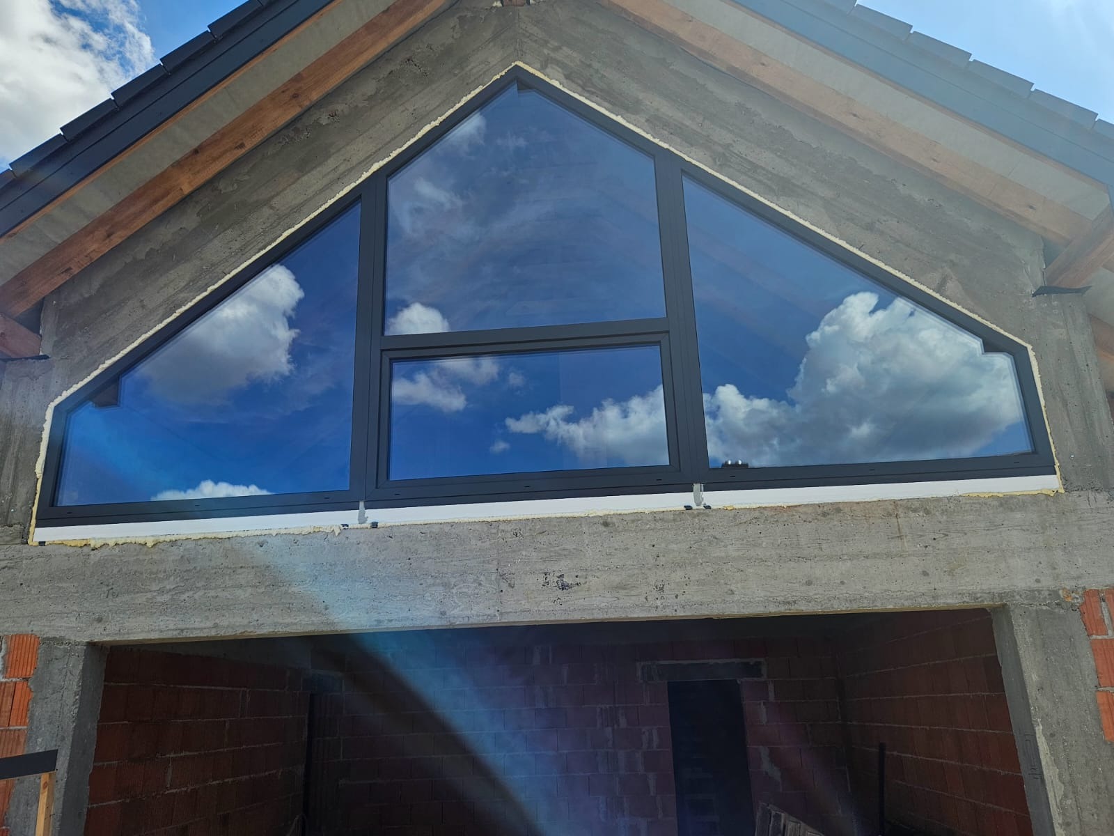 Trójkątne okno z czarną ramą, odbijające błękitne niebo z chmurami, zamontowane w surowej, betonowej konstrukcji budynku w trakcie budowy.