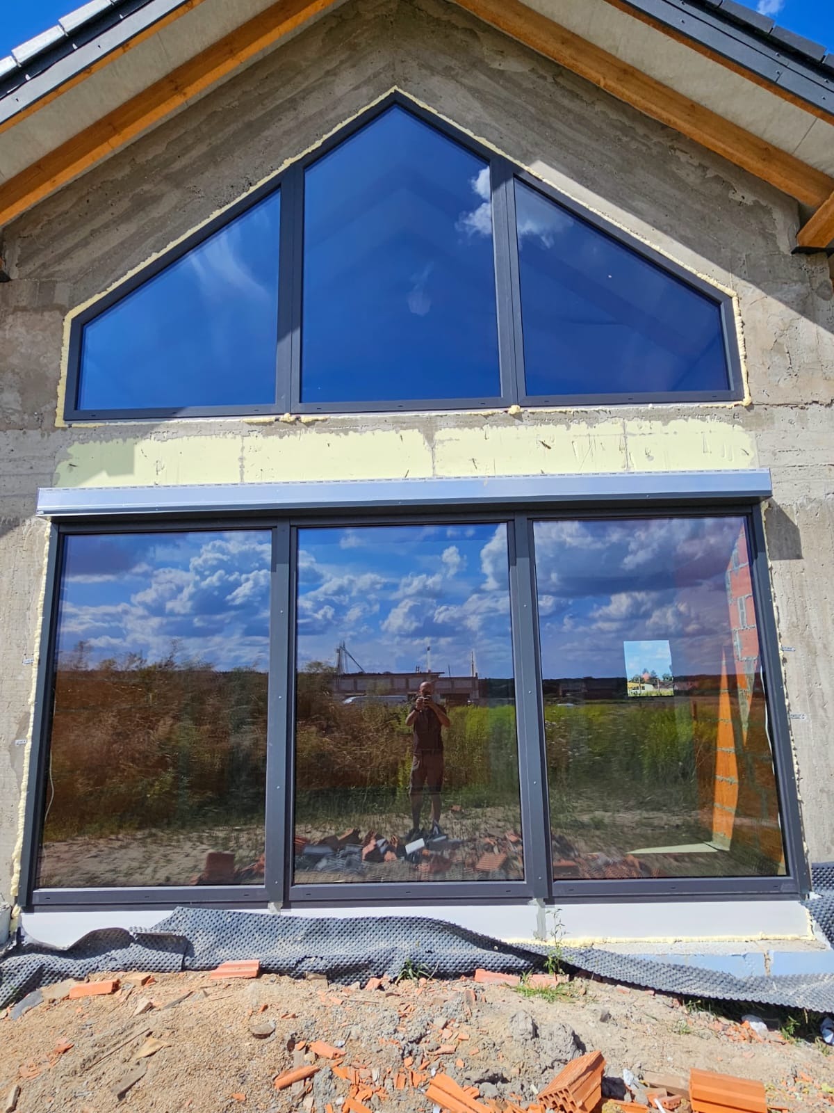 Nowe okna PCV w surowym budynku z szarą elewacją. Trójkątne okno na górze i duże okno na dole z roletą. Widoczne odbicie fotografa w oknach.