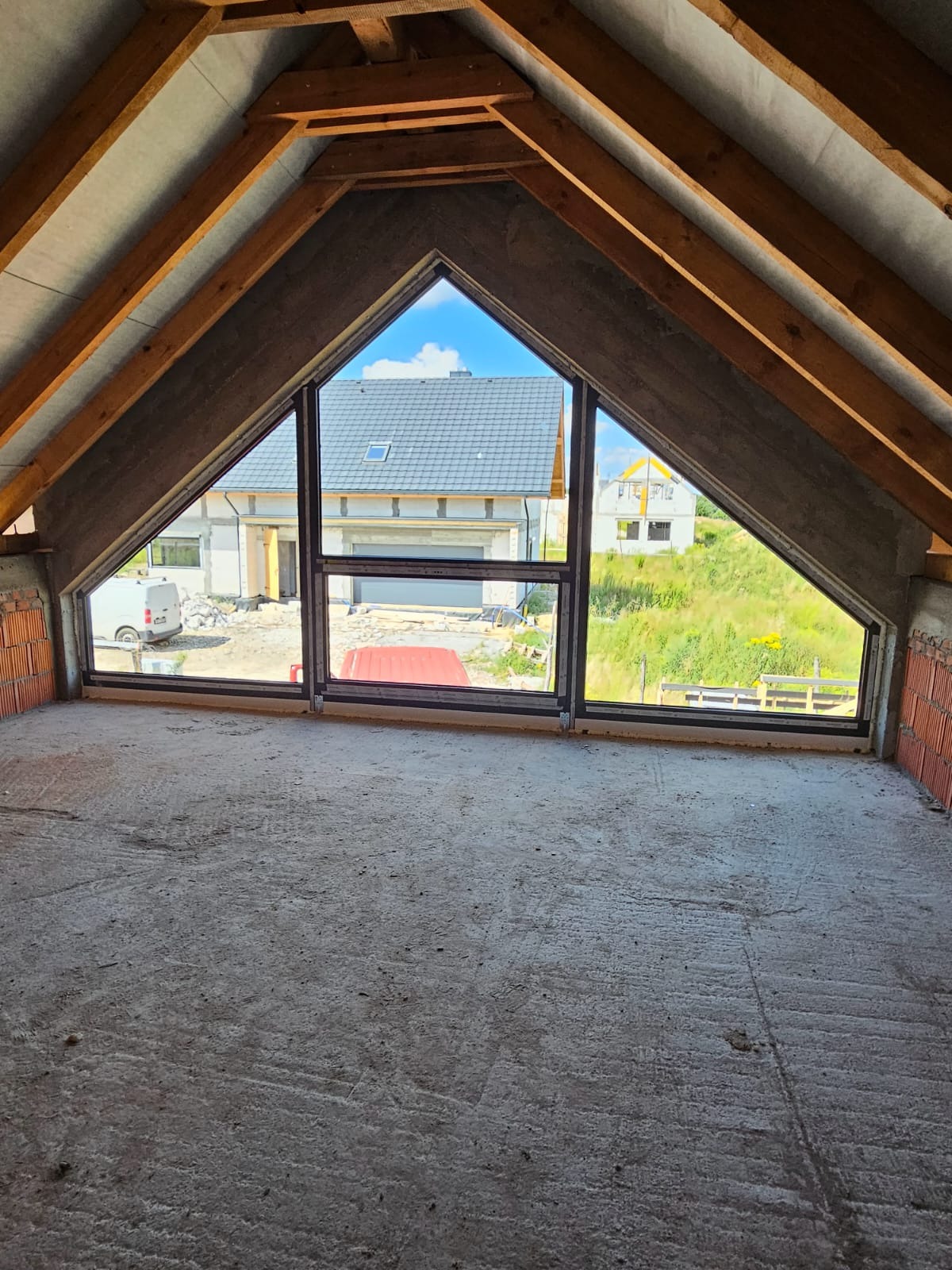 Surowy strych z oknami o nietypowym kształcie, widok na domy i zieleń. Okna z czarnymi ramami, surowy beton i cegła. Drewniana konstrukcja dachu.