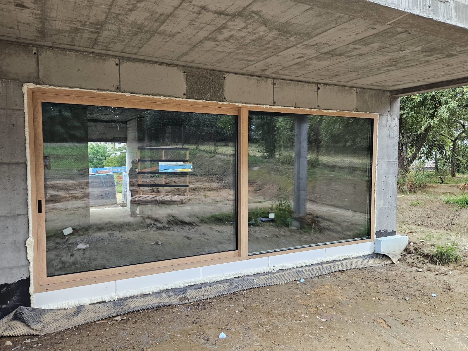 Montaż dużych, przesuwnych okien z drewnianą ramą w surowym stanie budynku. Widoczne pianka montażowa i folia hydroizolacyjna. Przeszklenia odbijają otoczenie.