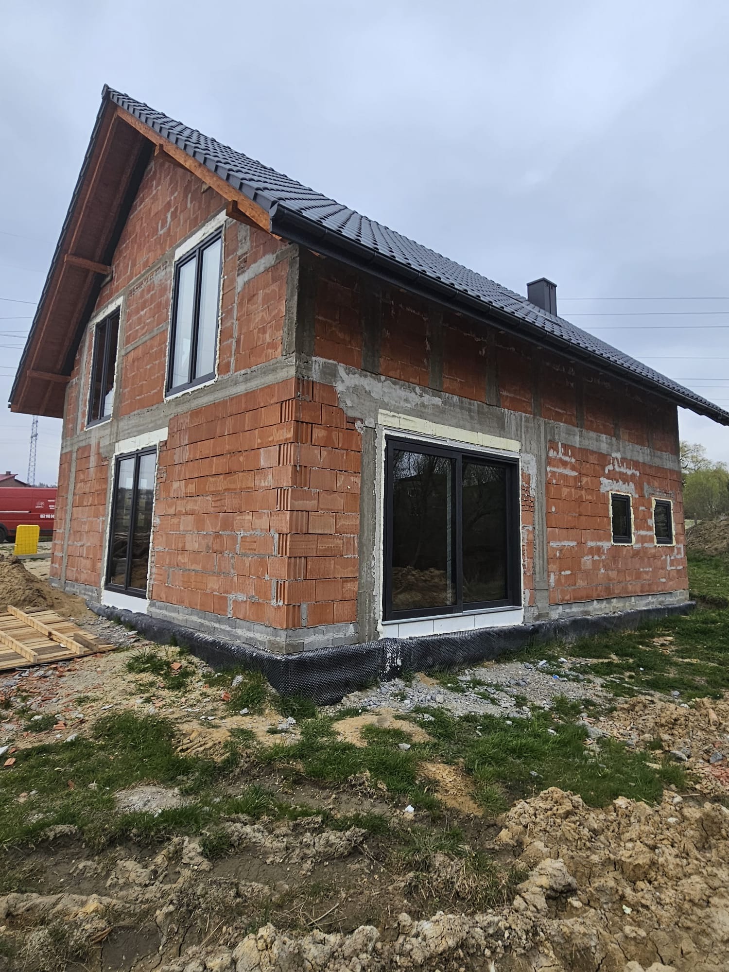 Surowy budynek z czerwonej cegły z czarnymi ramami okiennymi i ciemnym dachem, w trakcie budowy, widok z zewnątrz w pochmurny dzień.
