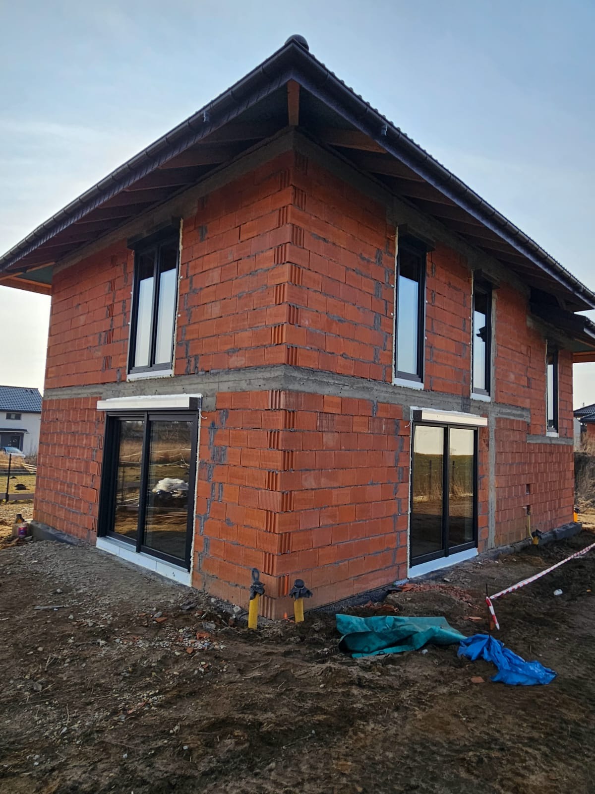 Surowy budynek z czerwonej cegły z zamontowanymi oknami i drzwiami przesuwnymi w czarnych ramach. Widoczne elementy instalacji na zewnątrz budynku.