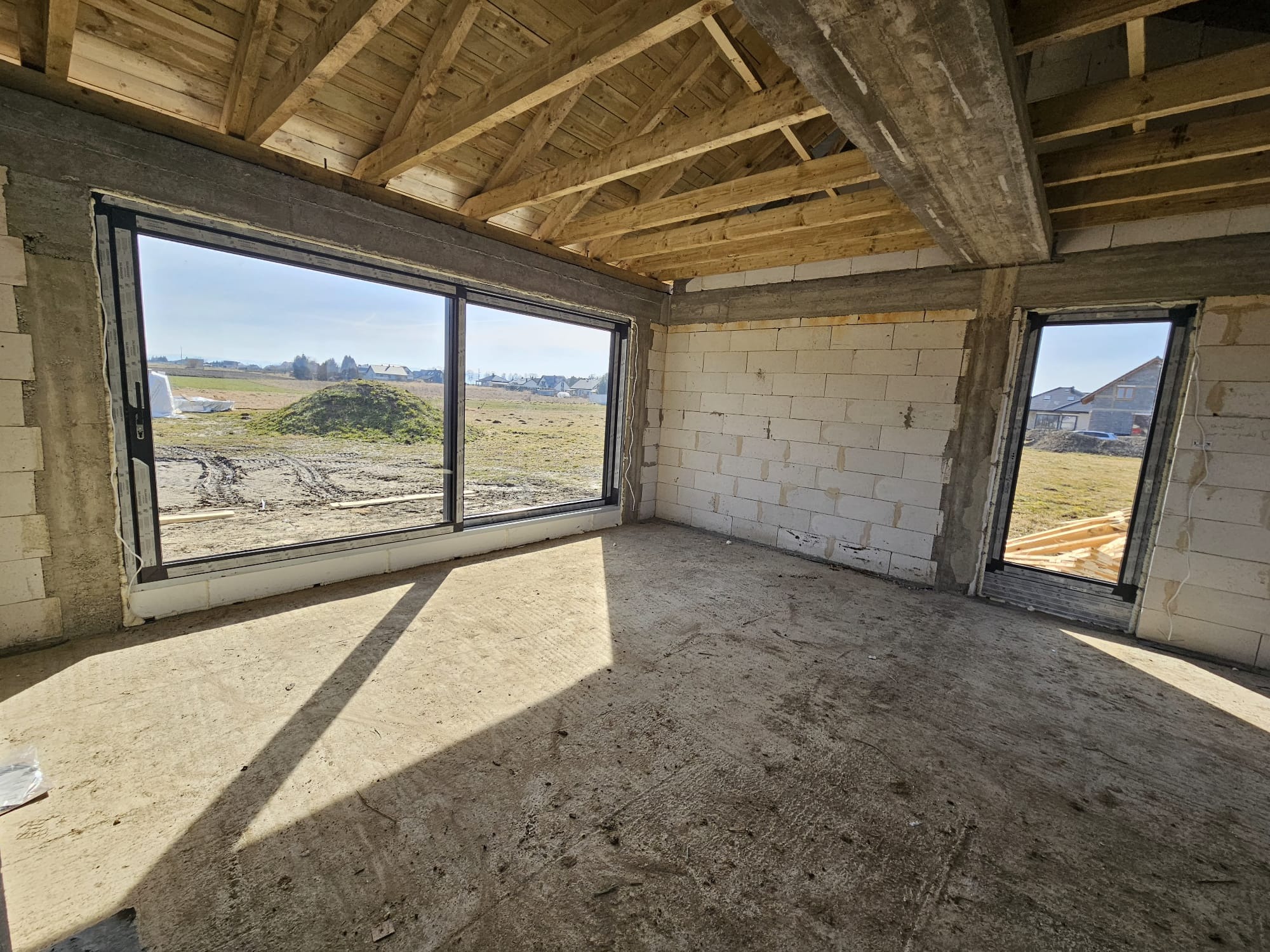 Surowy stan wnętrza z dużym oknem przesuwnym i mniejszym oknem, widok na zewnątrz, drewniana konstrukcja dachu, ściany z białych bloczków, betonowa podłoga w promieniach słońca.