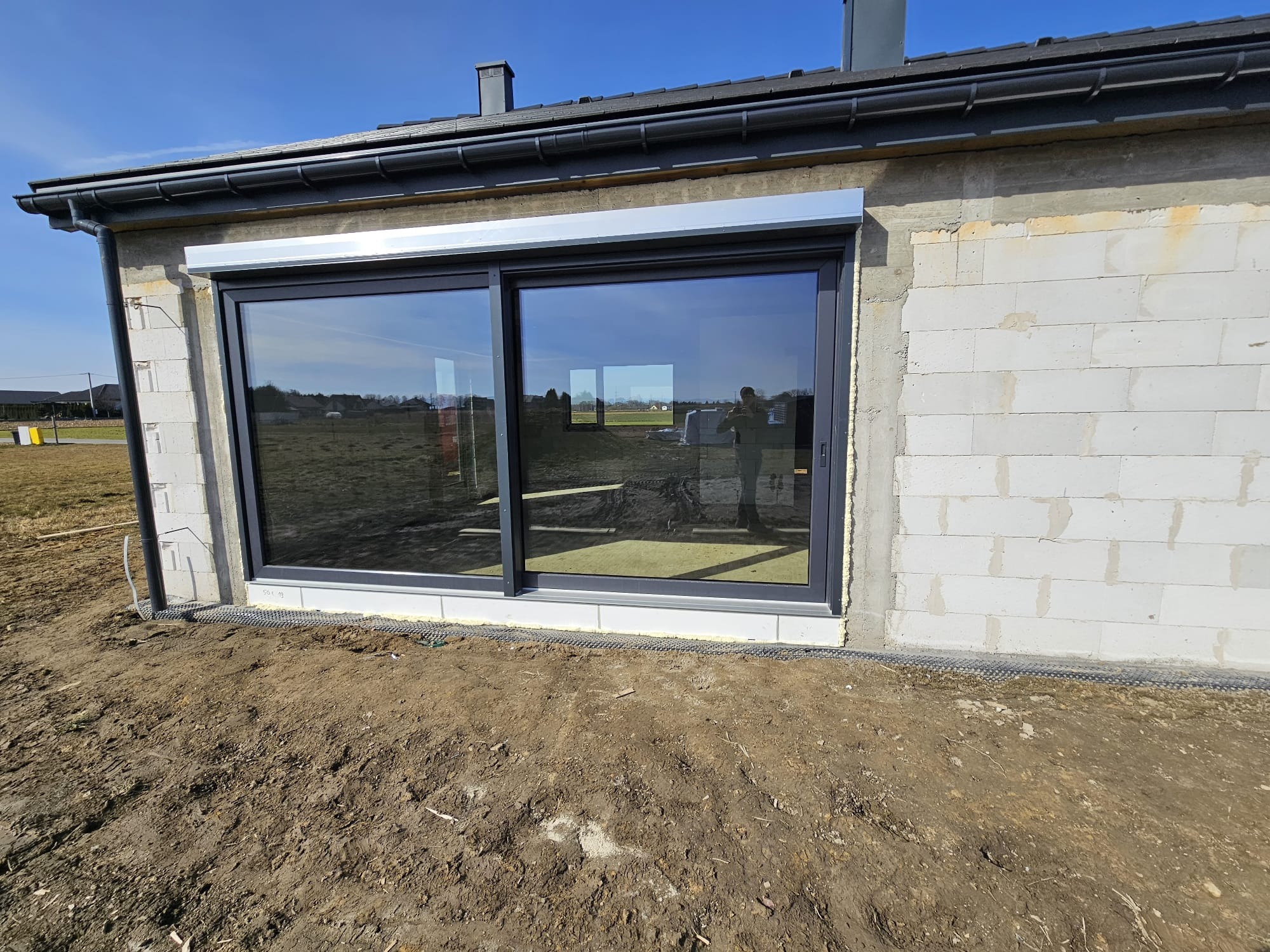 Zamontowane, grafitowe drzwi przesuwne z roletą zewnętrzną w surowym budynku z betonu komórkowego. W szybie odbija się sylwetka człowieka.