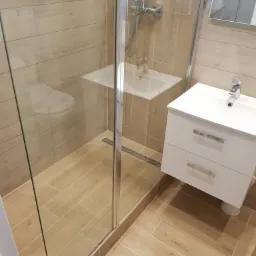 Nowoczesna łazienka z kabiną prysznicową ze szklanymi drzwiami, umywalką z szafką w kolorze białym i drewnopodobnymi płytkami na ścianach i podłodze, widoczny odpływ liniowy.