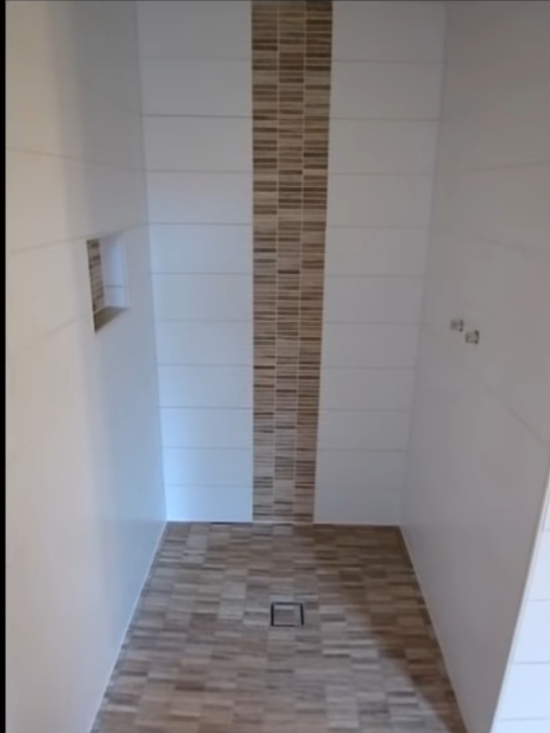 Nowoczesna kabina prysznicowa z białymi płytkami ułożonymi poziomo i dekoracyjnym pasem mozaiki w odcieniach brązu, z odpływem liniowym w podłodze wyłożonej płytkami imitującymi drewno.