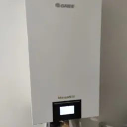 Biała pompa ciepła Gree Versati III zamontowana na ścianie, widoczne rury przyłączeniowe i wyświetlacz.