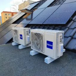 Dwa białe klimatyzatory Haier na dachu z panelami słonecznymi, zamontowane na białych wspornikach, na tle budynku mieszkalnego i błękitnego nieba.